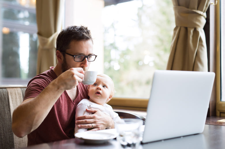 Man holding baby sitting in front of laptop drinking from a coffee mug